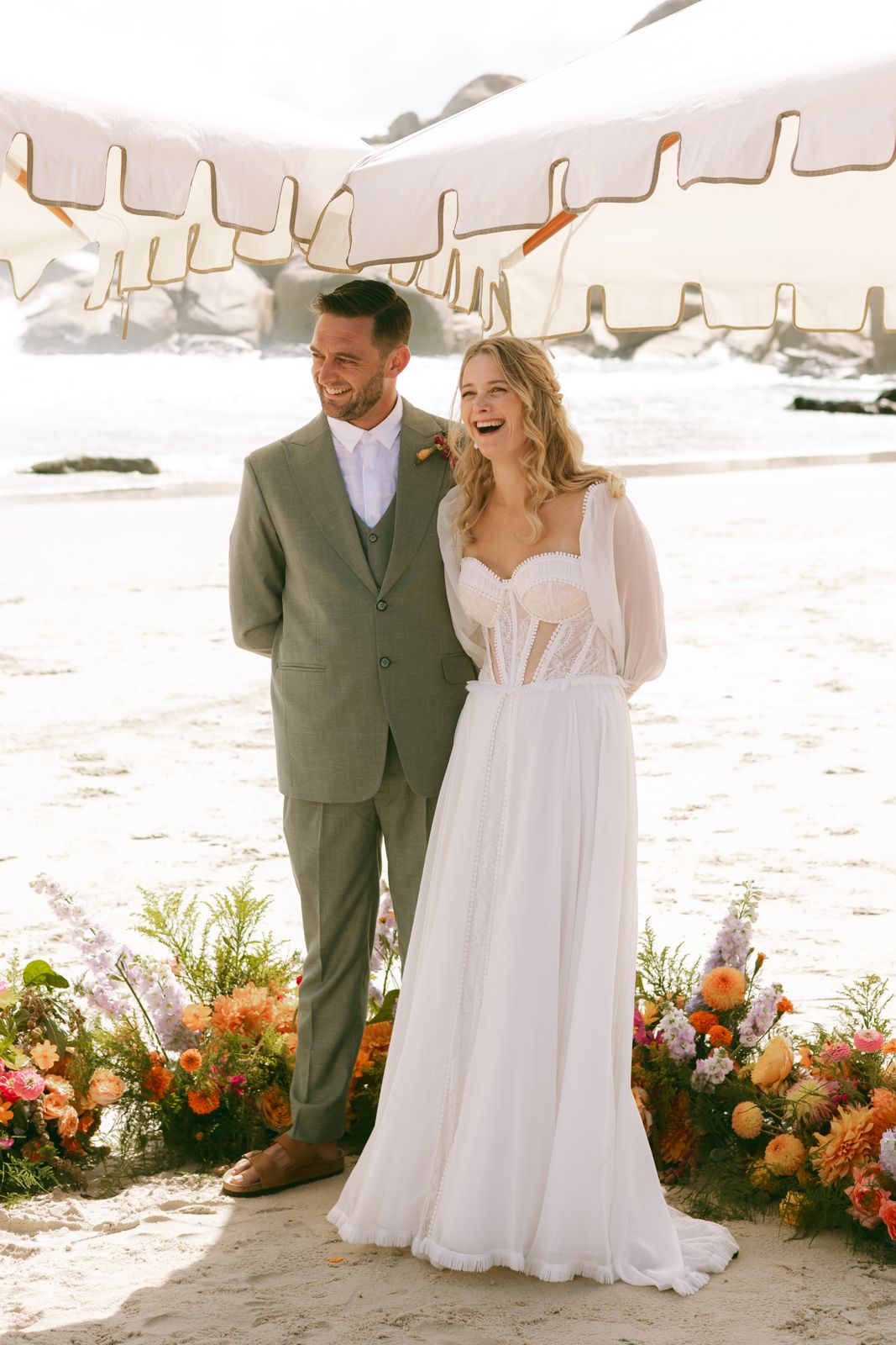 Bride and groom laughing beneath beach umbrellas