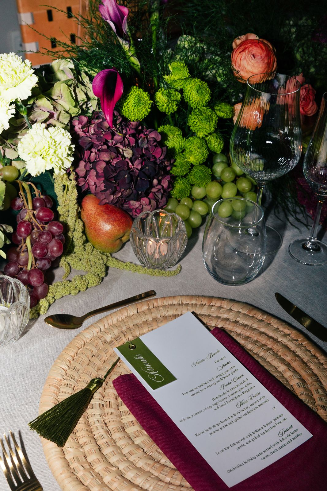 Menu card with tassel on a woven placemat beside a fruit and flower centrepiece