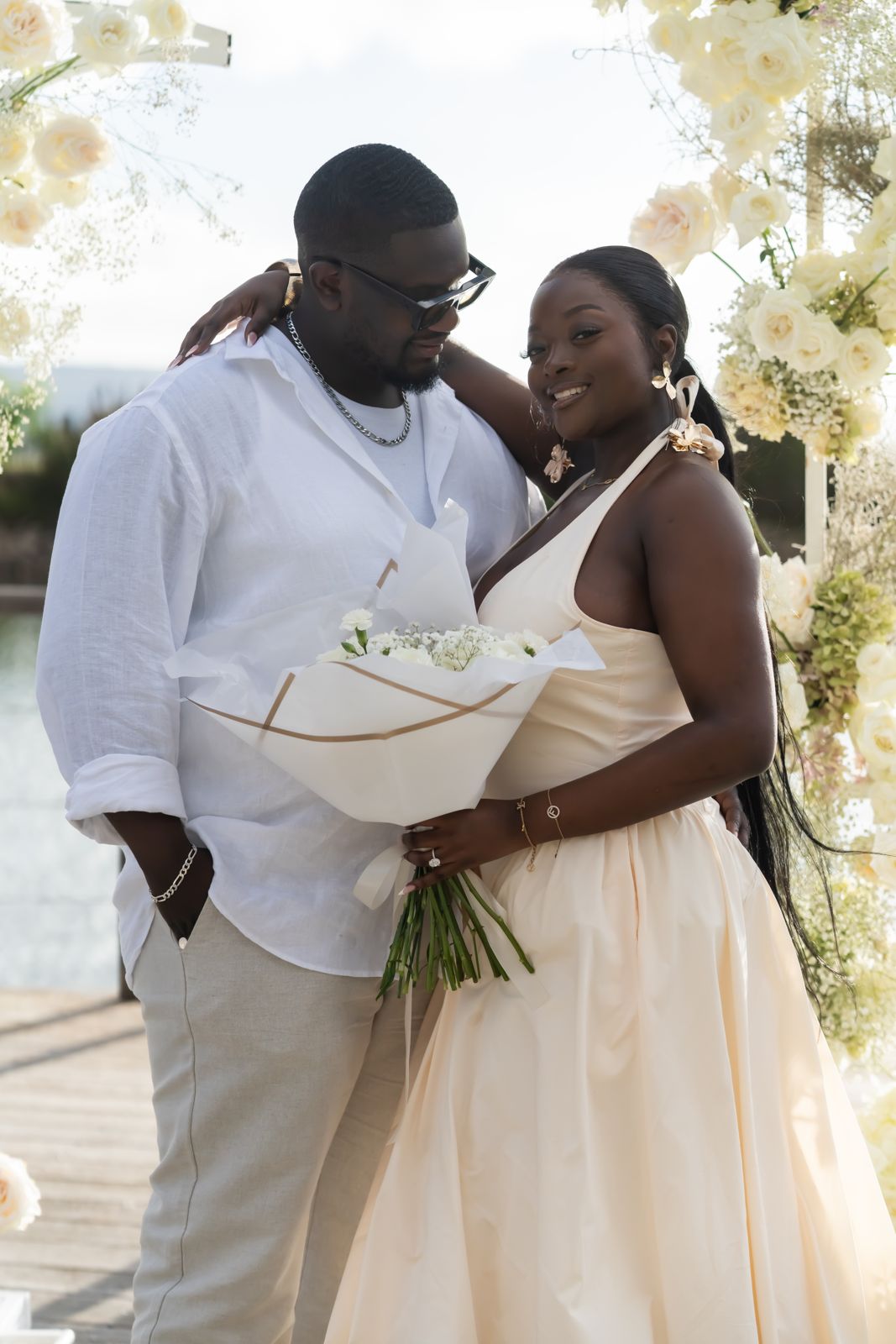 Newly engaged couple with a bouquet beneath a white rose arch