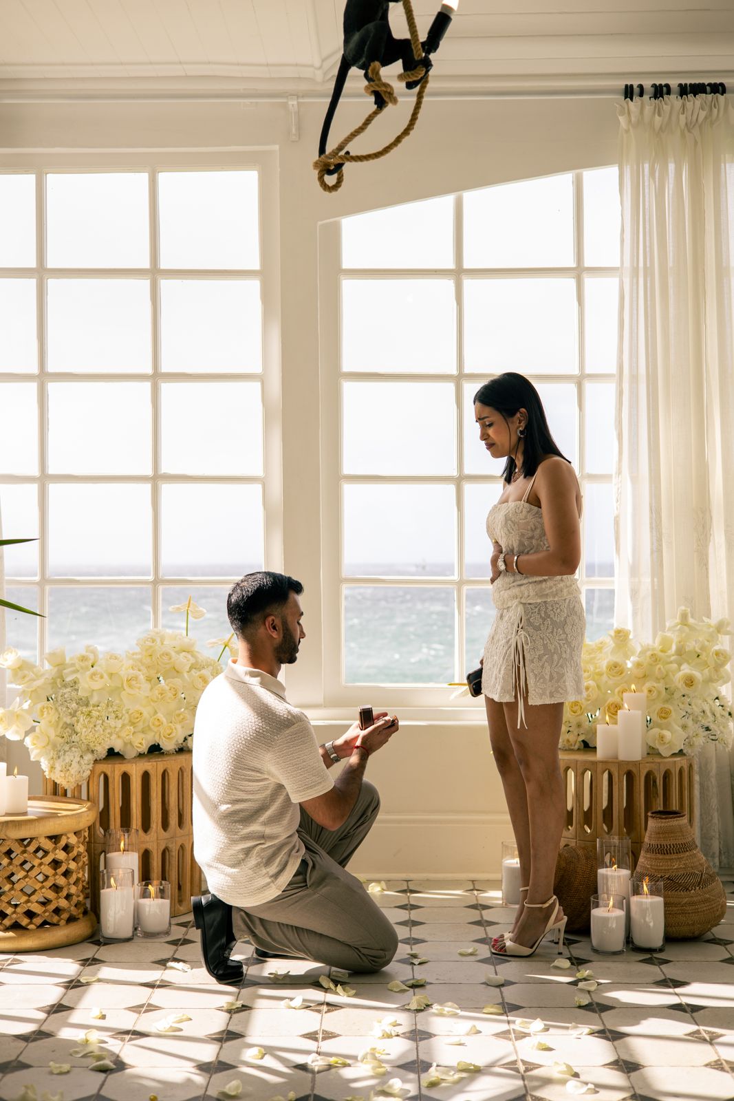 Proposal by tall windows overlooking the sea, with white roses and candles